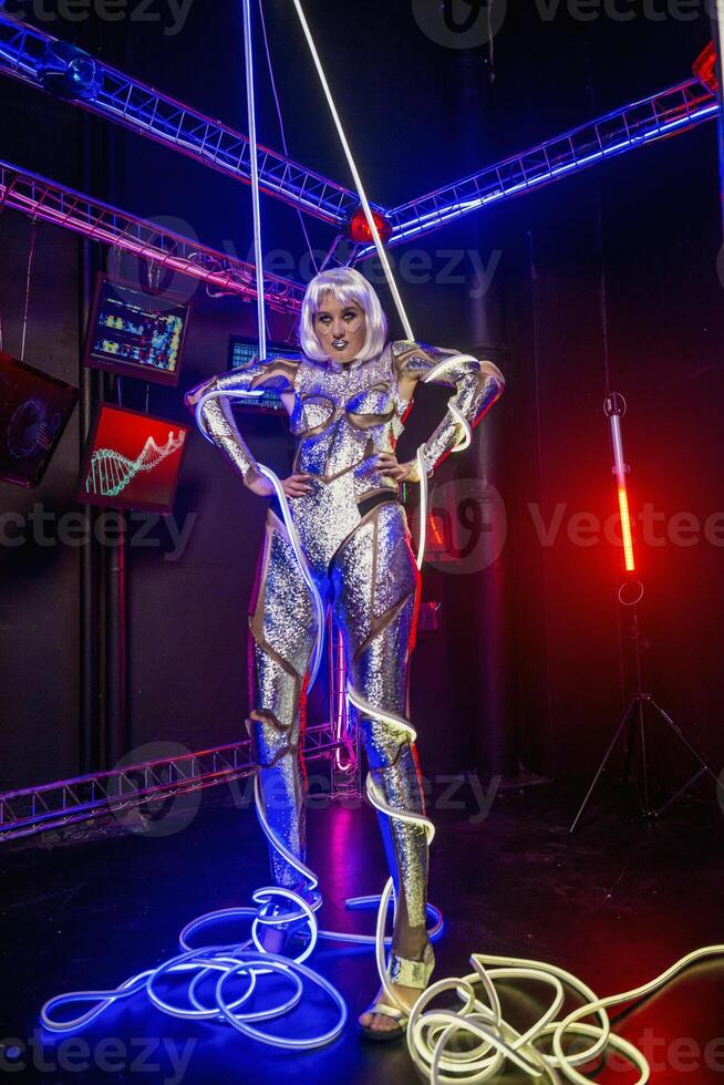 Blonde as a cyber robot. Girl in shining silver armor in neon. Blonde with fantastic makeup and image in dark key and colored light, posing in cyberspace. Her body is wrapped in glowing neon wires photo