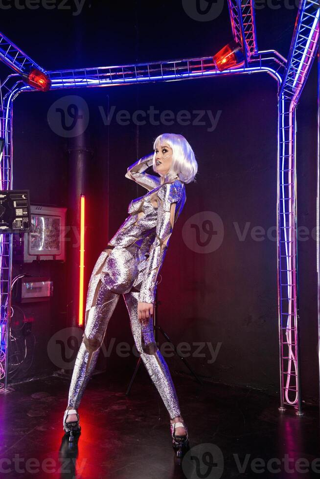 Blonde as cyber robot. Girl in shining silver armor in neon. Blonde with fantastic makeup and image in dark key and colored light, posing in cyberspace. She stands in a glowing cube, general plan photo