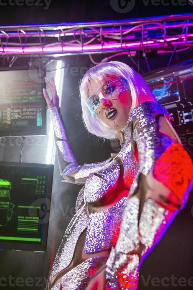 close-up of blonde as cyber robot. Girl in shining silver armor in neon, in glowing glasses. screens glow behind her. Blonde with fantastic makeup, posing in cyberspace. red light illuminates her photo