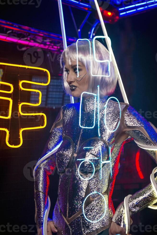 Blonde as a cyber robot. Girl in shining silver armor in neon. Blonde with fantastic makeup and image in dark key and colored light, posing in cyberspace. Letters are projected on her photo