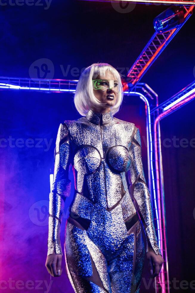 Blonde as a cyber robot. Girl in shining silver armor in neon. She walks in a glowing cube, smoke around. Blonde with fantastic makeup and image in a dark key and colored light, posing in cyberspace photo