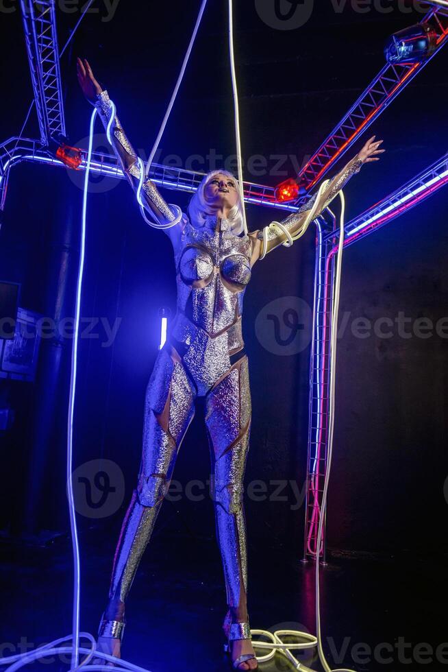 Blonde as a cyber robot. Girl in shining silver armor in neon. Blonde with fantastic makeup and image in dark key and colored light, posing in cyberspace. wires around hands, general plan photo