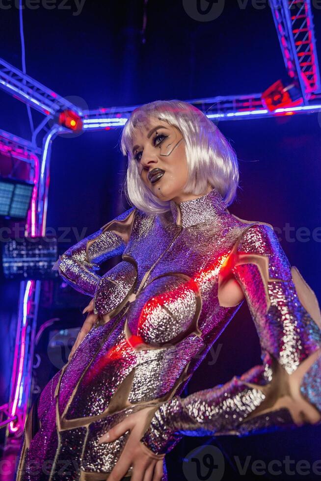 Blonde as a cyber robot. Girl in shining silver armor in neon. Blonde with fantastic makeup and image in dark key and colored light, posing in cyberspace. Wires glow around her photo
