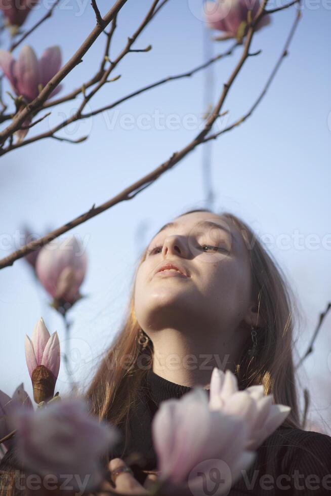 young blonde girl and pink blooming magnolia. happy girl standing near blooming tree in park. she has long straight hair and dark coat. she looks at sky, angle from below photo