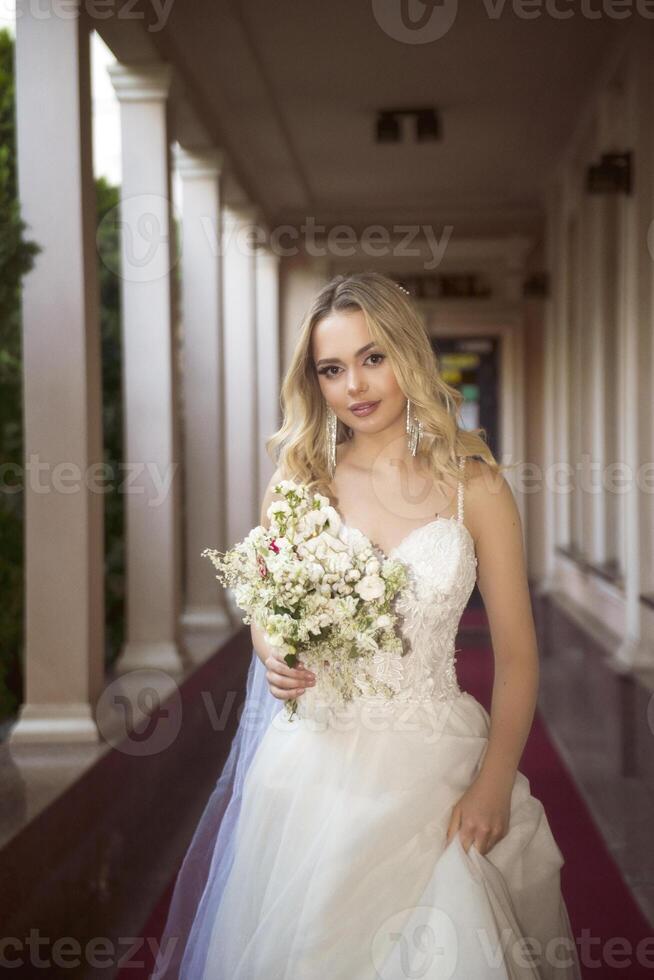 beautiful blonde bride walks along the open gallery of the hotel with a bouquet. long wedding dress, with open shoulders. wavy blonde hair, open shoulders, she smiles and looks at the camera photo