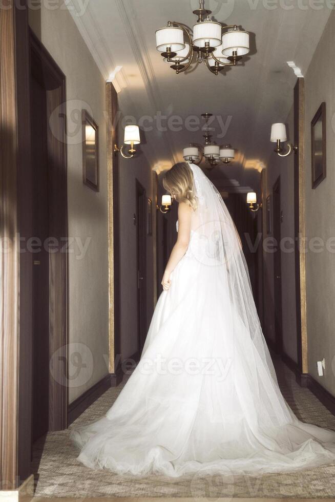 beautiful blonde bride with veil in hotel hallway in fashionable dress, back view. elegant bride in lace dress, with bare shoulders and stylish wave hairstyle and makeup.dress with train and long veil photo