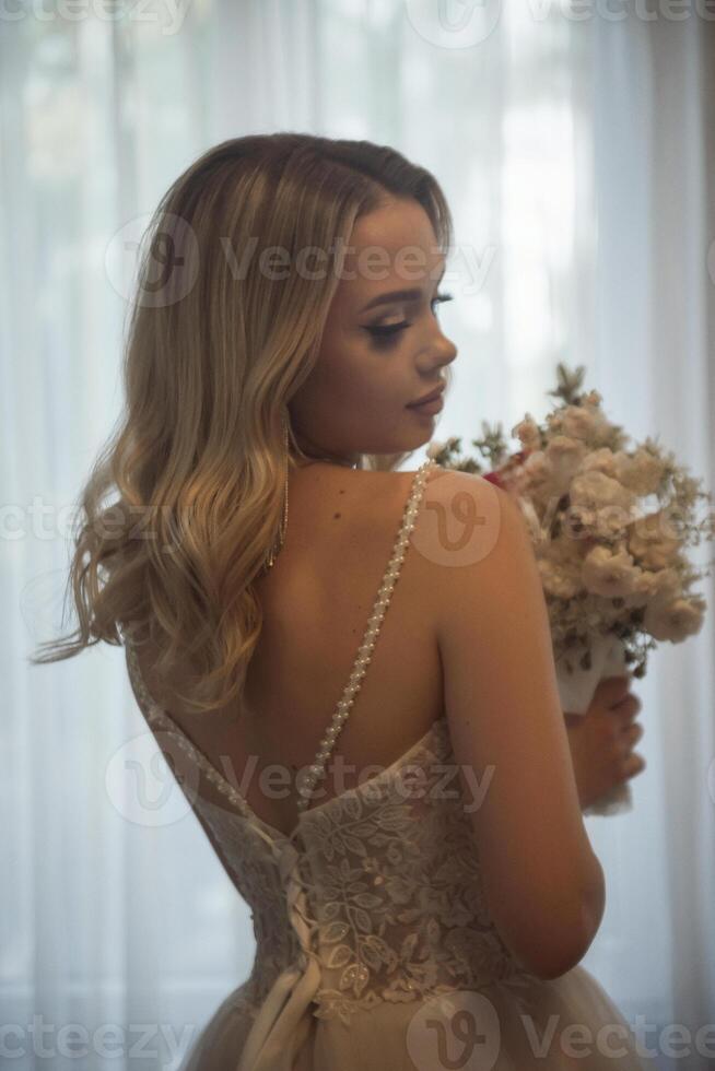close-up of the bride's silhouette in a long wedding dress from the back with a bouquet in her hands. beautiful young blonde stands against the backdrop of a hotel window photo