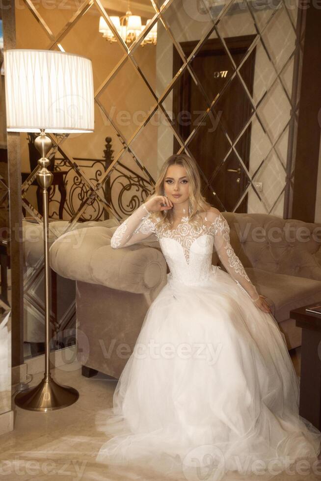 beautiful blonde bride in a long white dress sits in a hotel, against the background of a mirror wall. fashionable dress with long sleeves made of chiffon and lace. light hair loose and stylishly styled. photo