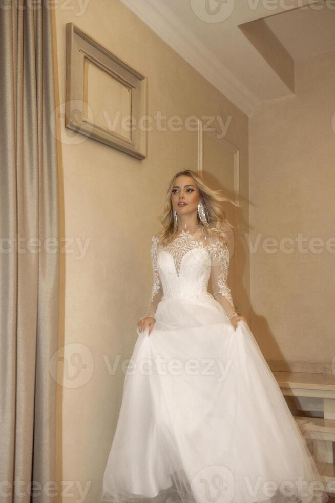 beautiful blonde bride walking down the stairs in the hotel. in a long wedding dress, with a corset made of lace and chiffon. she has wavy blonde hair, beautiful earrings. she smiles photo