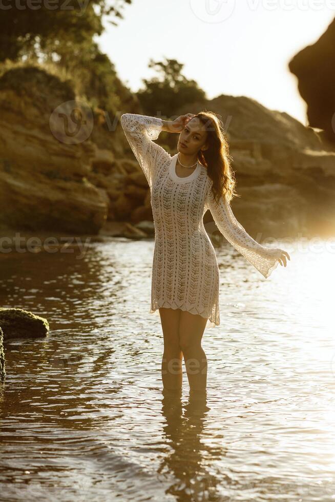 Beautiful young woman posing on the beach in summer. girl standing in backlight. her dress glows in the sun, in white knitted dress with pearls on her neck, in water against the backdrop of a rock photo