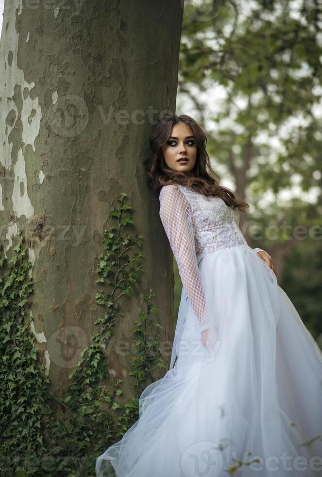 young beautiful girl bride in elegant wedding dress in park. blooming trees in spring. long dress made of lace and chiffon. girl thoughtful. concept of european wedding fashion. photo