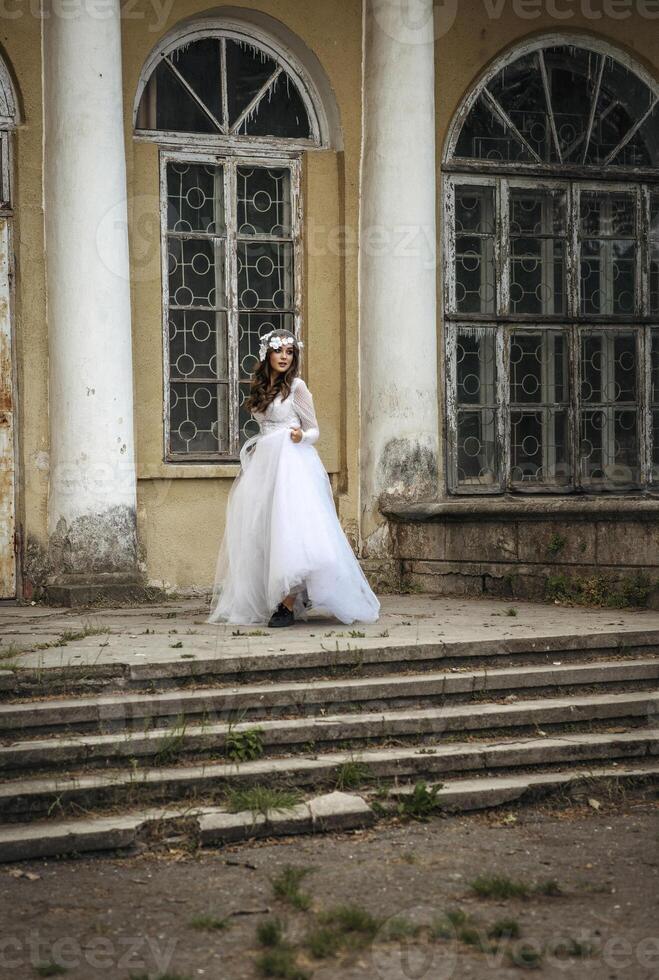 young girl bride in a white dress in a spring forest on a wedding day, beautiful brunette in a long stylish plan among huge trees. hair veil with flowers, against background of 19th century building photo