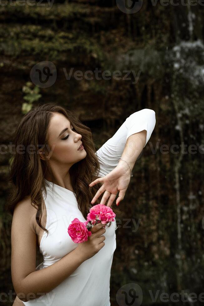 charming girl with roses in her hands in nature against the backdrop of rocks. with fashionable makeup and dark wavy hair posing in nature. in a white bodysuit with a slanted neckline. in the background a waterfall photo