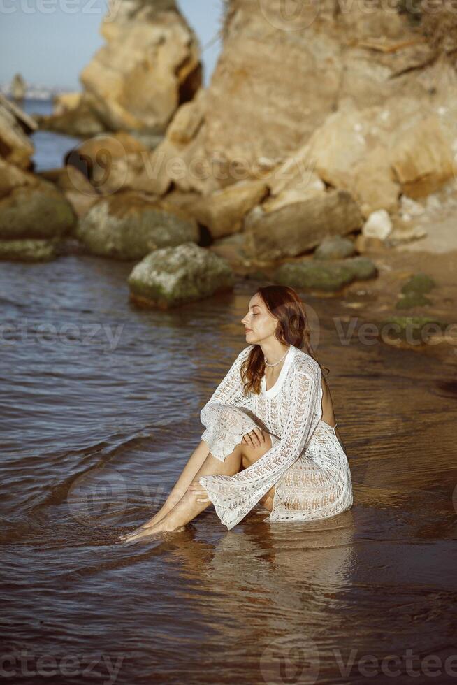 beautiful slim young girl sits on the sea beach, in the sea. around her the sea surface, she is dressed in a white knitted dress, against the background of large rocks. concept of a summer sea morning photo