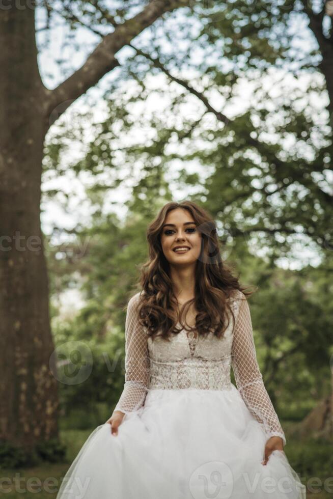 young girl bride in a white dress in a spring forest on a wedding day. beautiful brunette in a long stylish plan among huge trees. girl smiling and looking at camera. dress made of lace and chiffon photo