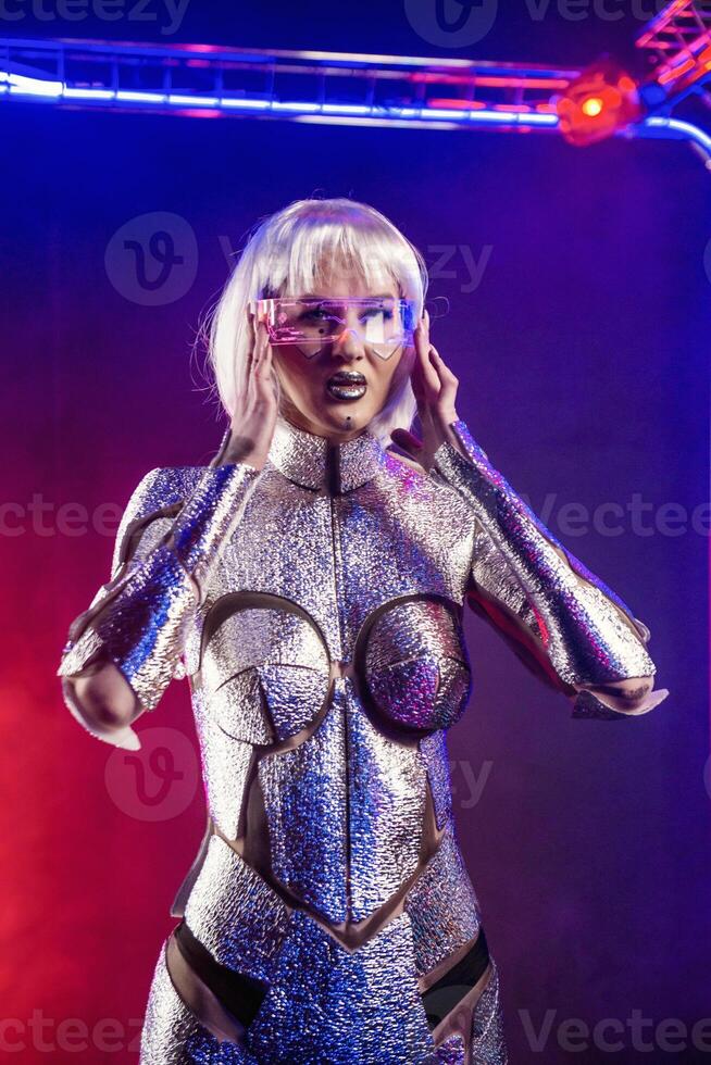 Blonde as a cyber robot. Girl in shining silver armor in neon. She stands in a glowing cube, she has transparent glasses. Blonde with fantastic makeup and image in a dark key and colored light photo
