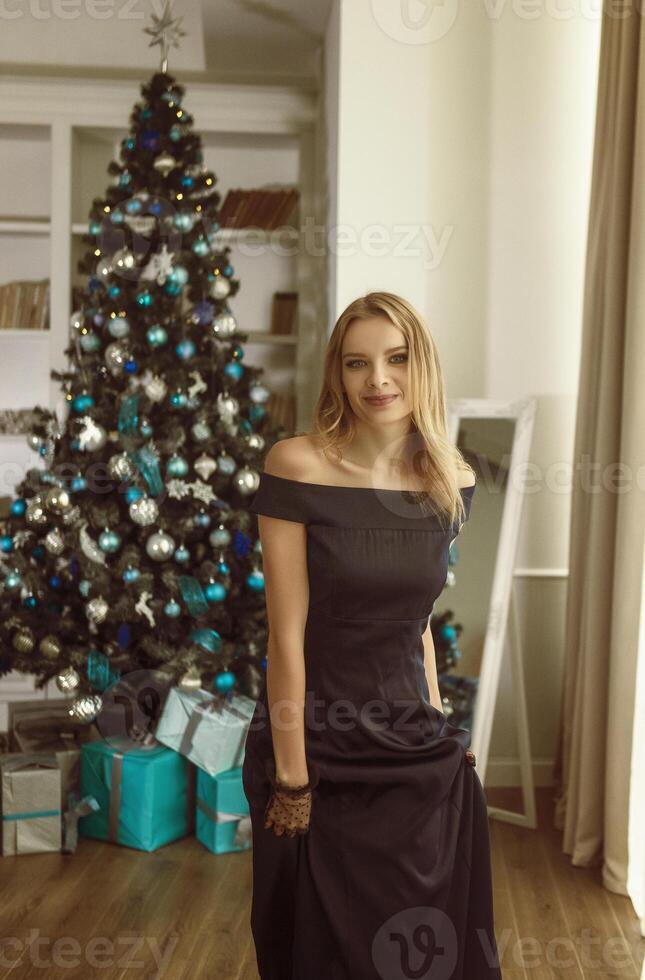beautiful young blonde in an evening dress poses against the background of a Christmas tree. near the tree there are gifts in turquoise boxes. concept of Christmas and New Year. looks at the camera photo