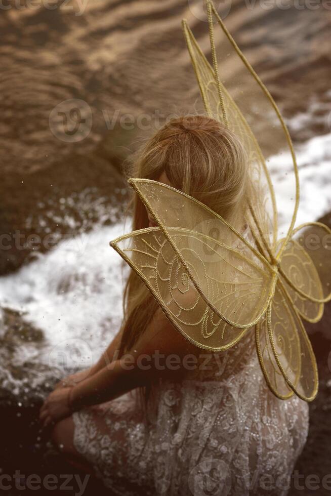 Cute blonde in fairy costume with wings and lace, transparent white dress sits in front of waterfall. Fairy wings are golden, made of transparent chiffon, decorated with embroidery. Concept of magic, Top view photo