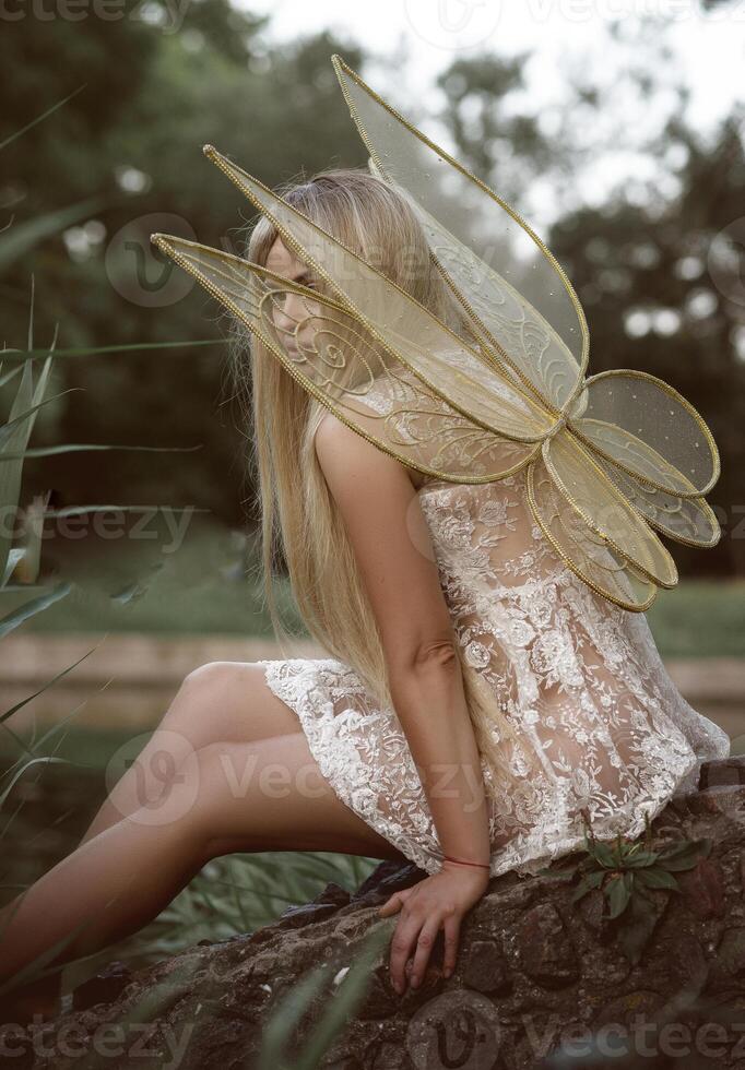 cute blonde girl in fairy costume with wings and lace, transparent white dress sits on the river bank. fairy wings of golden color, made of transparent chiffon, decorated with embroidery photo