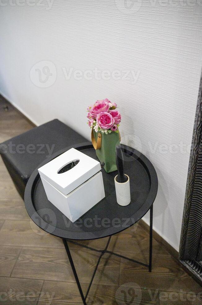 round black metal table next to it pouf. on the table are a box for napkins, decorative flowers, a brush for cleaning clothes. vertical, top view. on the floor are wood-like tiles. the walls are white photo