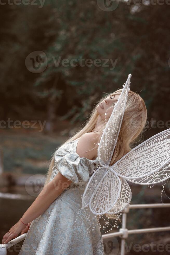 cute blonde girl in fairy costume with wings. girl in dress standing on river bank. fairy wings white, made of transparent chiffon, decorated with pearls. concept of magic and nature photo