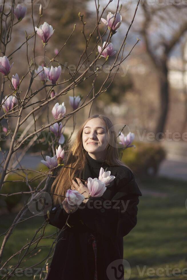 young blonde girl in the park near magnolia tree. happy girl laughing in the sun rays, pink flowers of the tree near her. she has long straight hair and dark coat photo