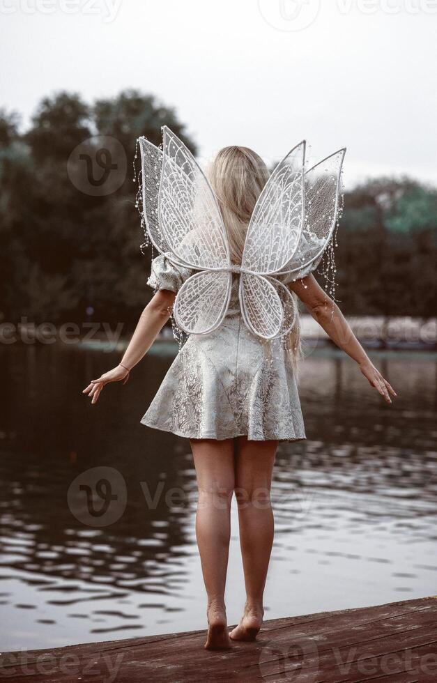 cute blonde girl in fairy costume with wings. girl in dress standing on river bank. fairy wings white, made of transparent chiffon, decorated with pearls. back view, full length. concept of magic and nature photo