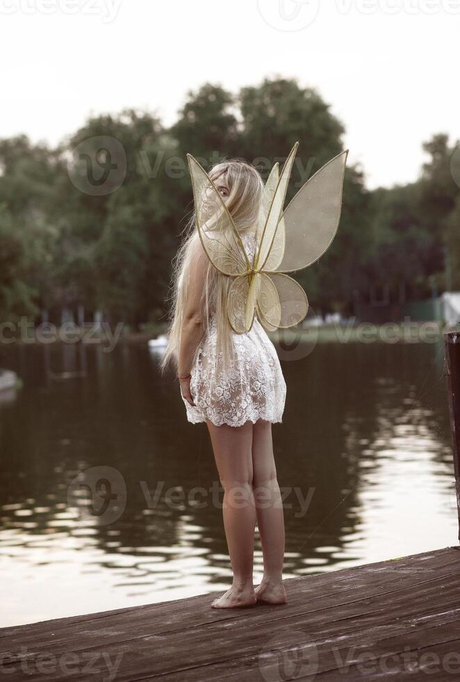 cute blonde girl in fairy costume with wings. girl on the river bank. golden fairy wings, made of transparent chiffon, decorated with embroidery. concept of magic and nature. back view, full length photo