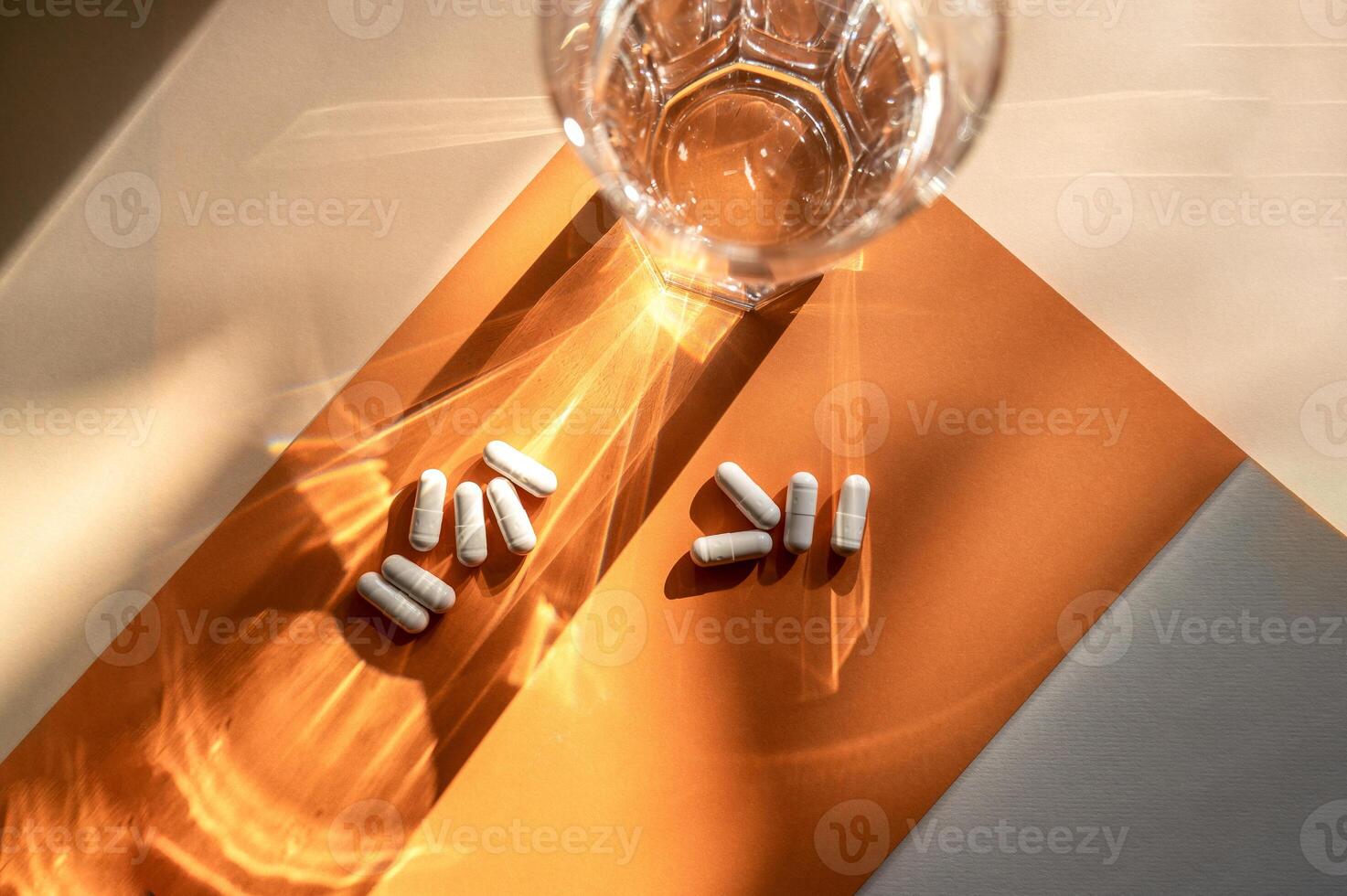 white medical capsules lie on the background, next to a transparent glass of water. A shadow from a glass of water falls on an orange background. top view, medical concept about medicine photo