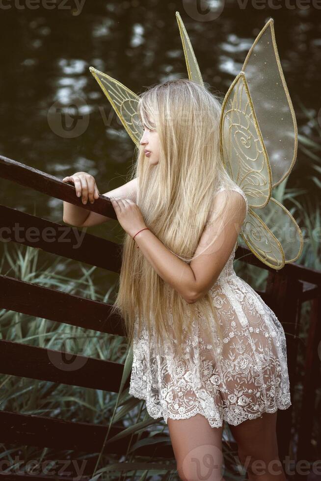 cute blonde girl in fairy costume with wings and lace white dress on the river bank. golden fairy wings, made of transparent chiffon, decorated with embroidery. concept of magic and nature photo
