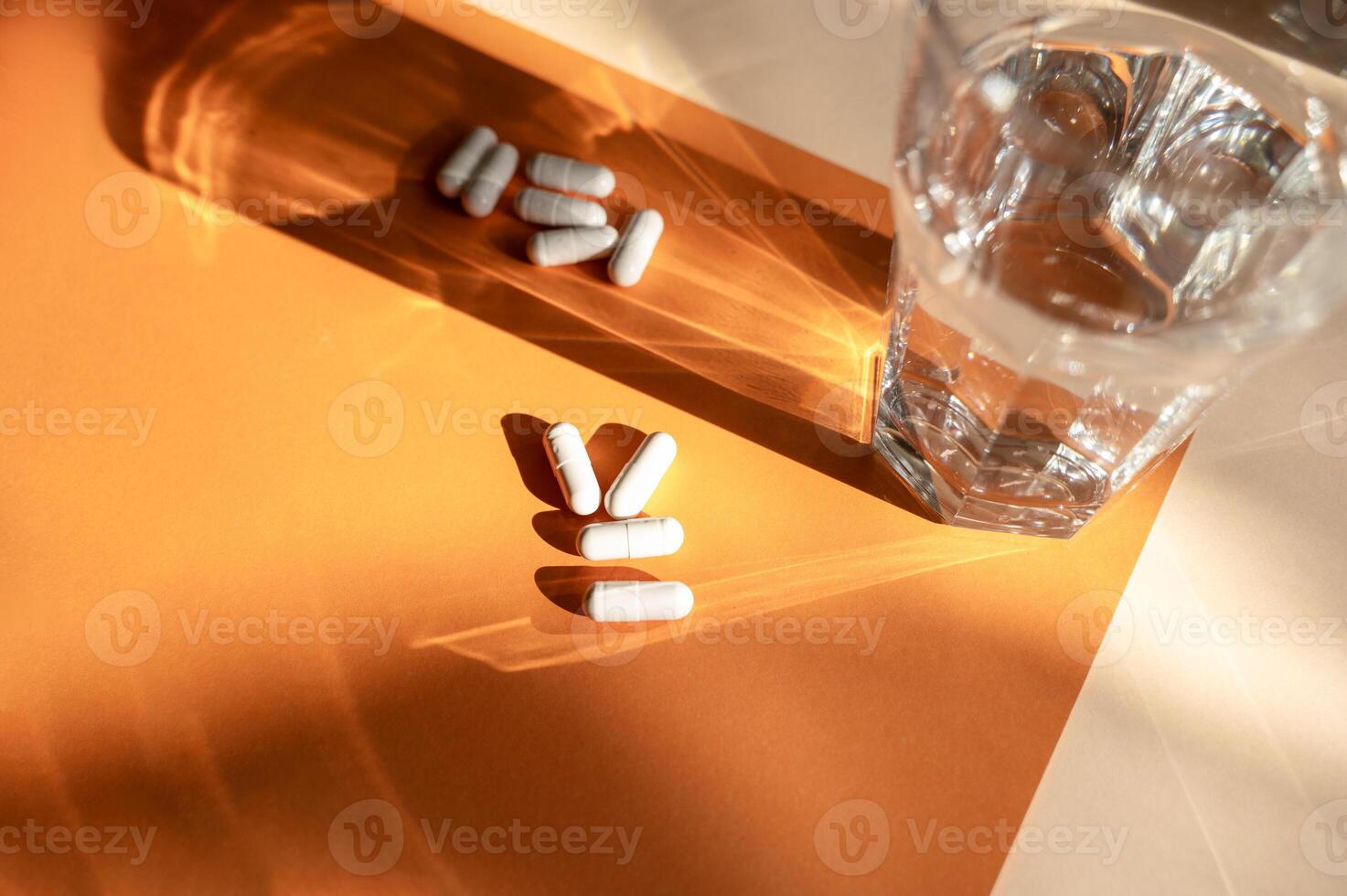 white medical capsules lie on the background, next to a transparent glass of water. A shadow from a glass of water falls on an orange background. top view. medical concept about medicine photo
