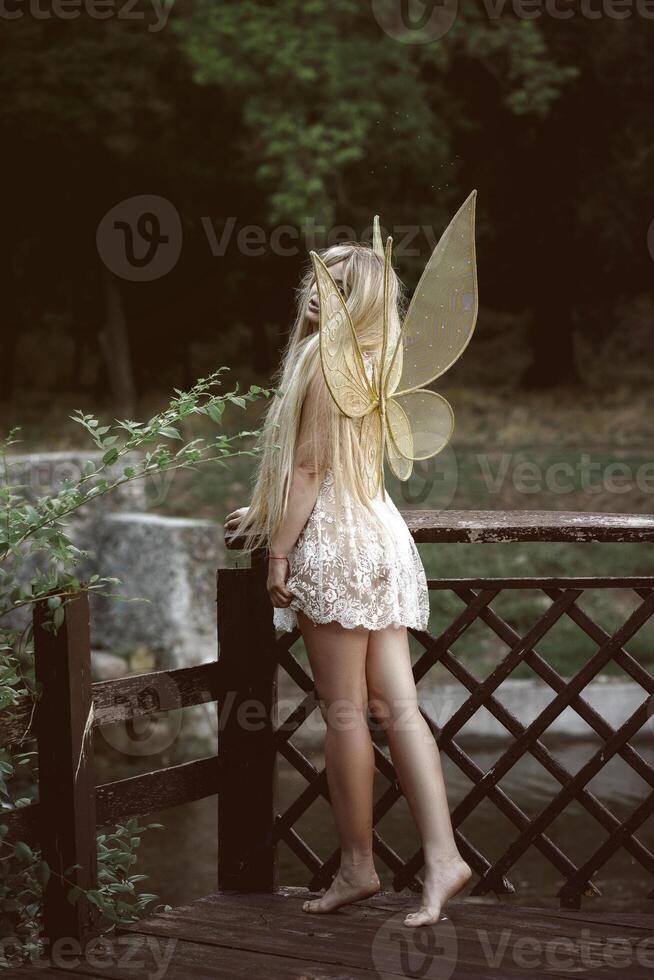 cute blonde girl in fairy costume with wings. girl in nature near the river. fairy wings of golden color, made of transparent chiffon, decorated with embroidery. concept of magic and nature. back view, full length photo