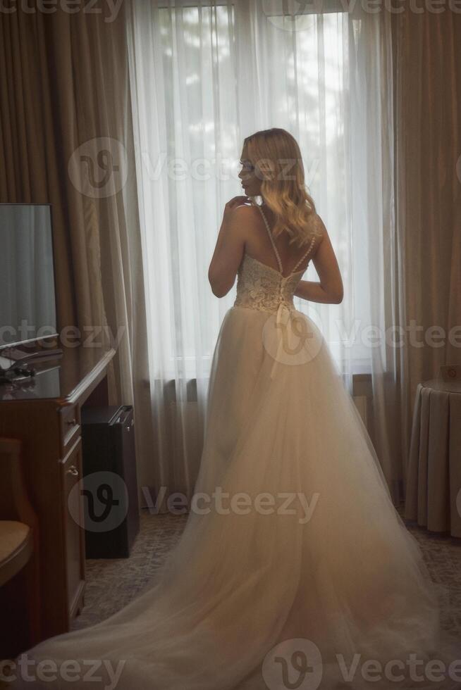 silhouette of a bride in a long wedding dress from the back. beautiful young blonde stands against the backdrop of a hotel window. long fashionable dress made of white chiffon photo