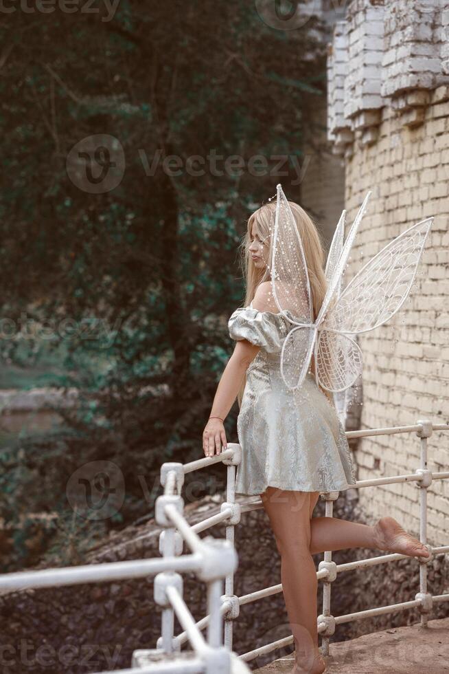 cute blonde girl in a fairy costume with wings. girl in a dress stands near the castle wall. fairy wings are white, made of transparent chiffon, decorated with artificial pearls. back view, full length. photo