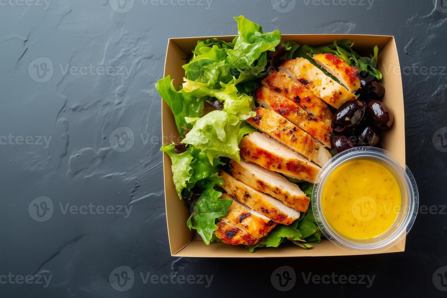 Grab-and-go salad with lettuce, grilled chicken, black olives, and lemon dressing in a cardboard box on a dark textured background. Fresh and healthy takeaway meal. . photo