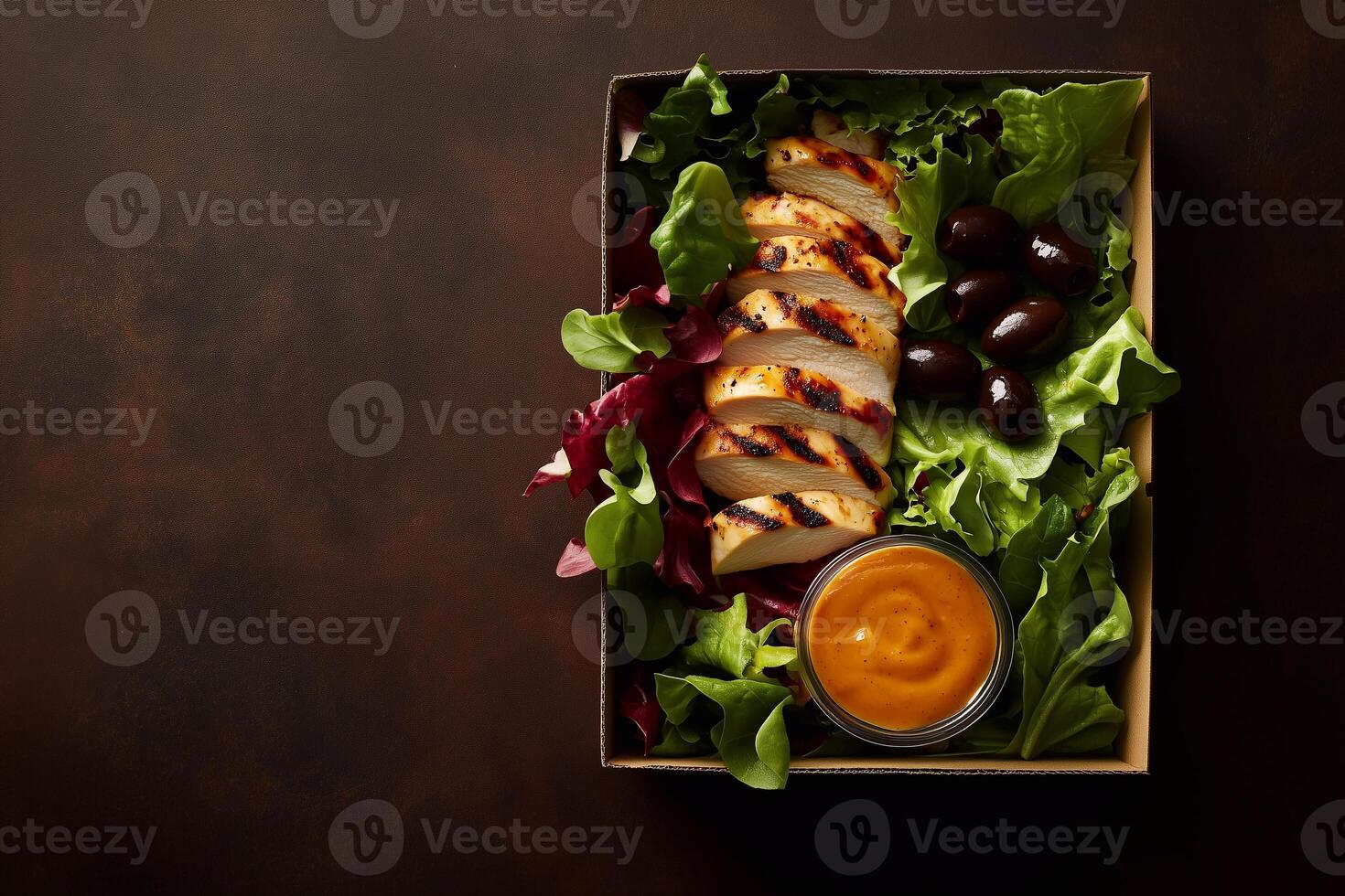 A grab-and-go salad in a rustic cardboard box with grilled chicken, fresh lettuce, black olives, and a side of golden dressing, set on a dark textured background. . photo