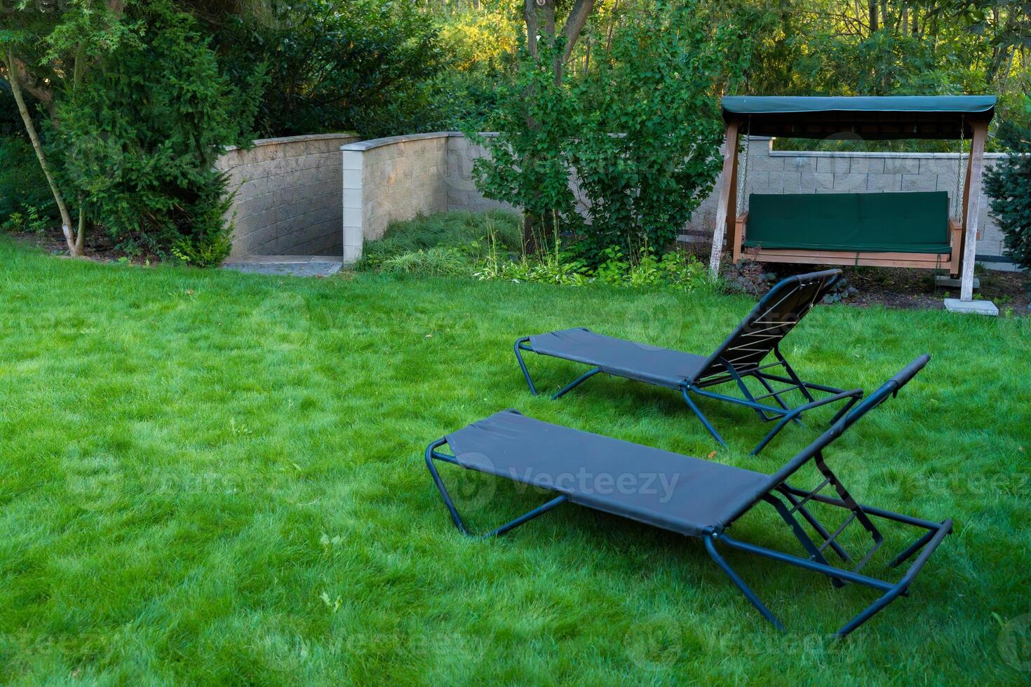 Peaceful backyard featuring two garden loungers and a swing bench set on a lush green lawn photo