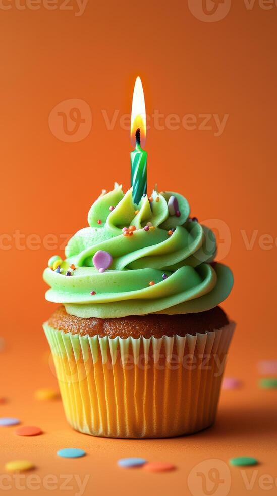 Colorful birthday cupcake with green frosting and a lit candle set against a vibrant orange background photo