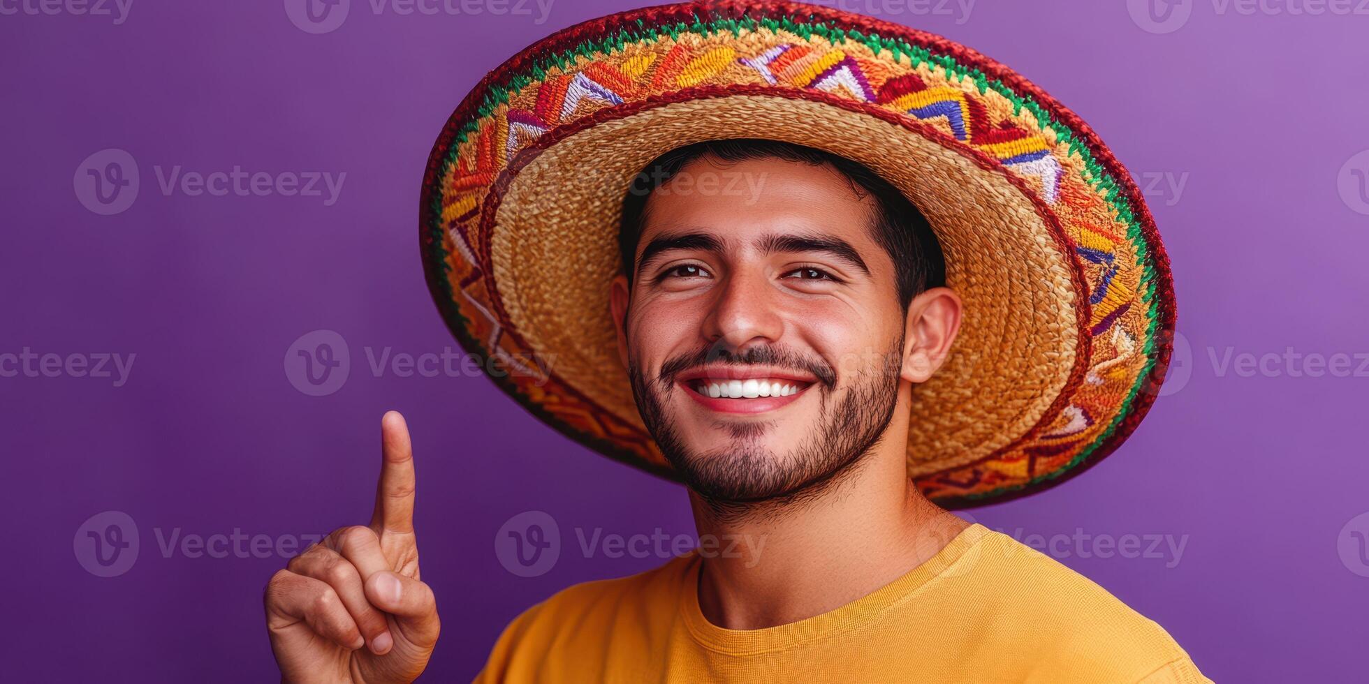 Young man wearing a colorful sombrero smiles and points in front of a vibrant purple background during a festive celebration photo