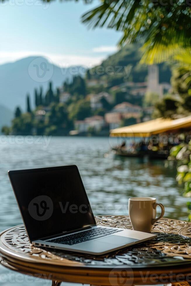 Laptop and coffee cup on a lakeside terrace with mountains and villas in the background photo