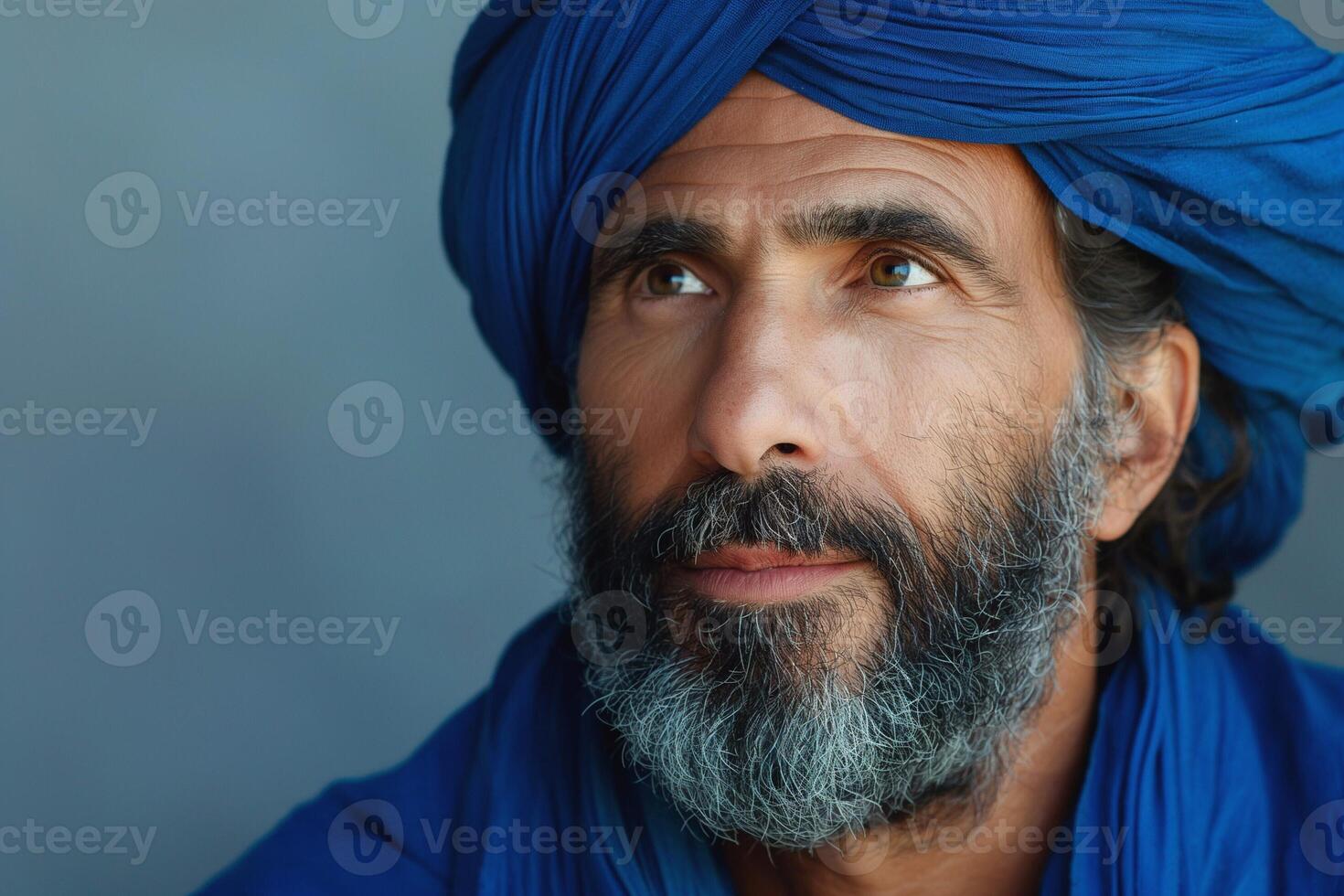 A contemplative Middle-Eastern man adorned in a blue turban and cloak gazes upward with a wise expression. photo