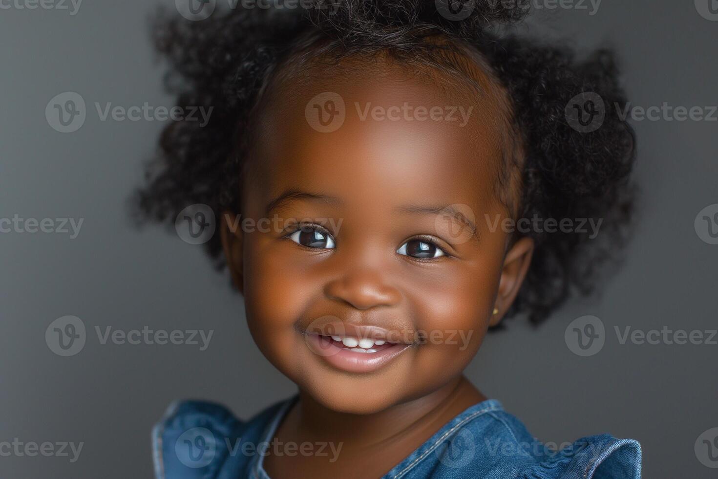 Adorable African American girl with curly hair beams with joy, wearing a denim outfit against a soft gray background. photo