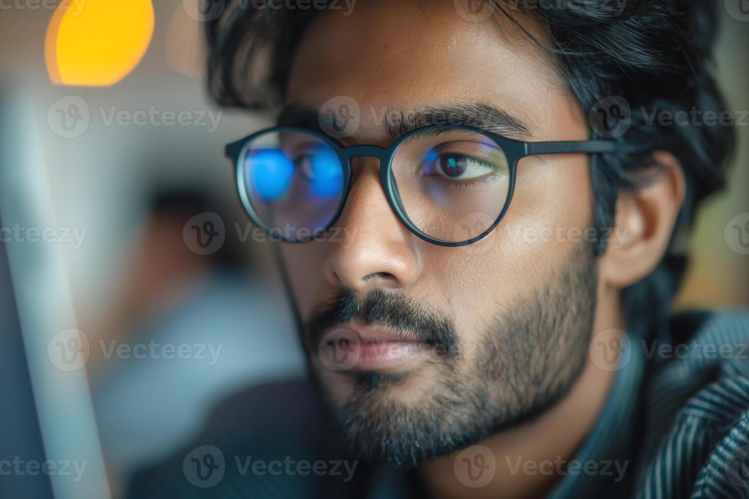 A focused South Asian man with glasses gazes intently at a computer screen, immersed in thought. photo