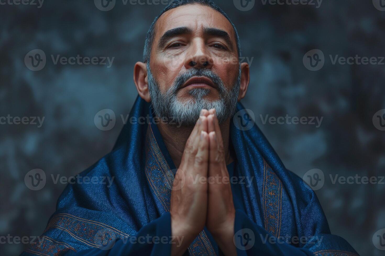 An elderly Middle-Eastern man with a gray beard, dressed in a dark shawl, prayerfully poses with his hands together. photo