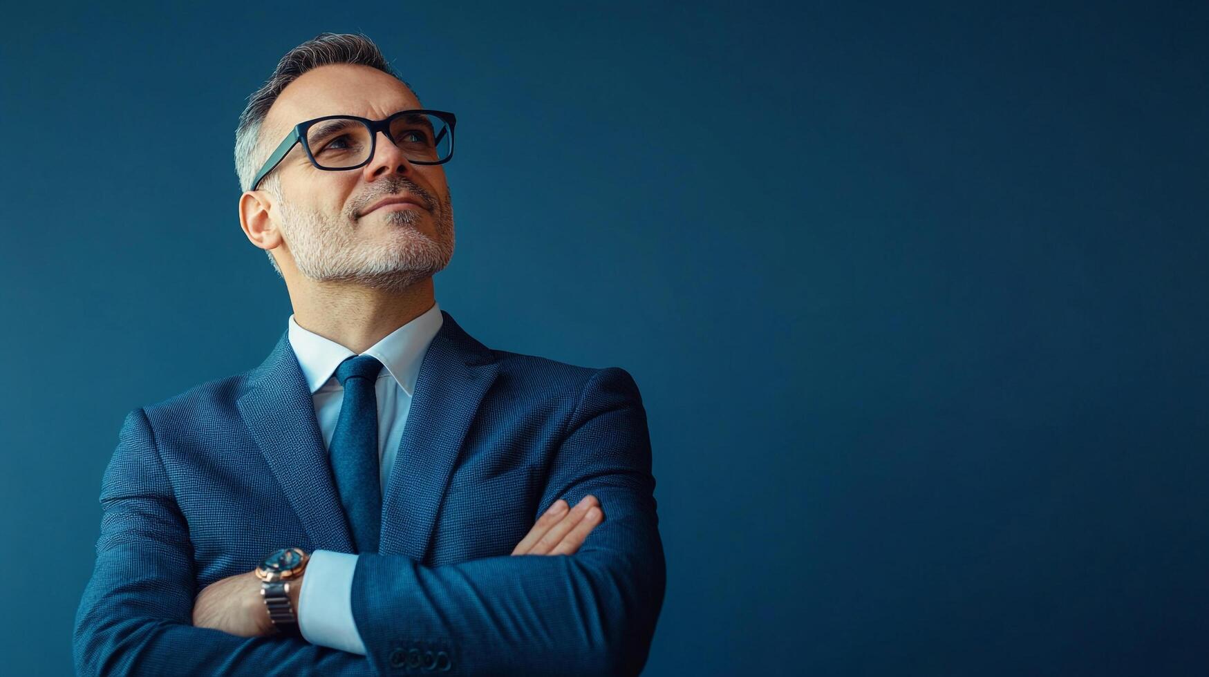 Confident businessman in a blue suit stands with arms crossed, looking upward with a hopeful expression against a solid backdrop. photo