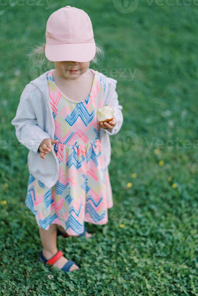 Little girl in a cap pulled down over her eyes with an apple walks on a green lawn photo