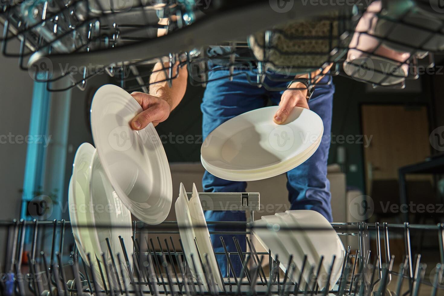 Man placing clean white plates into dishwasher, viewed from inside the appliance. Daily household routine in a modern kitchen photo