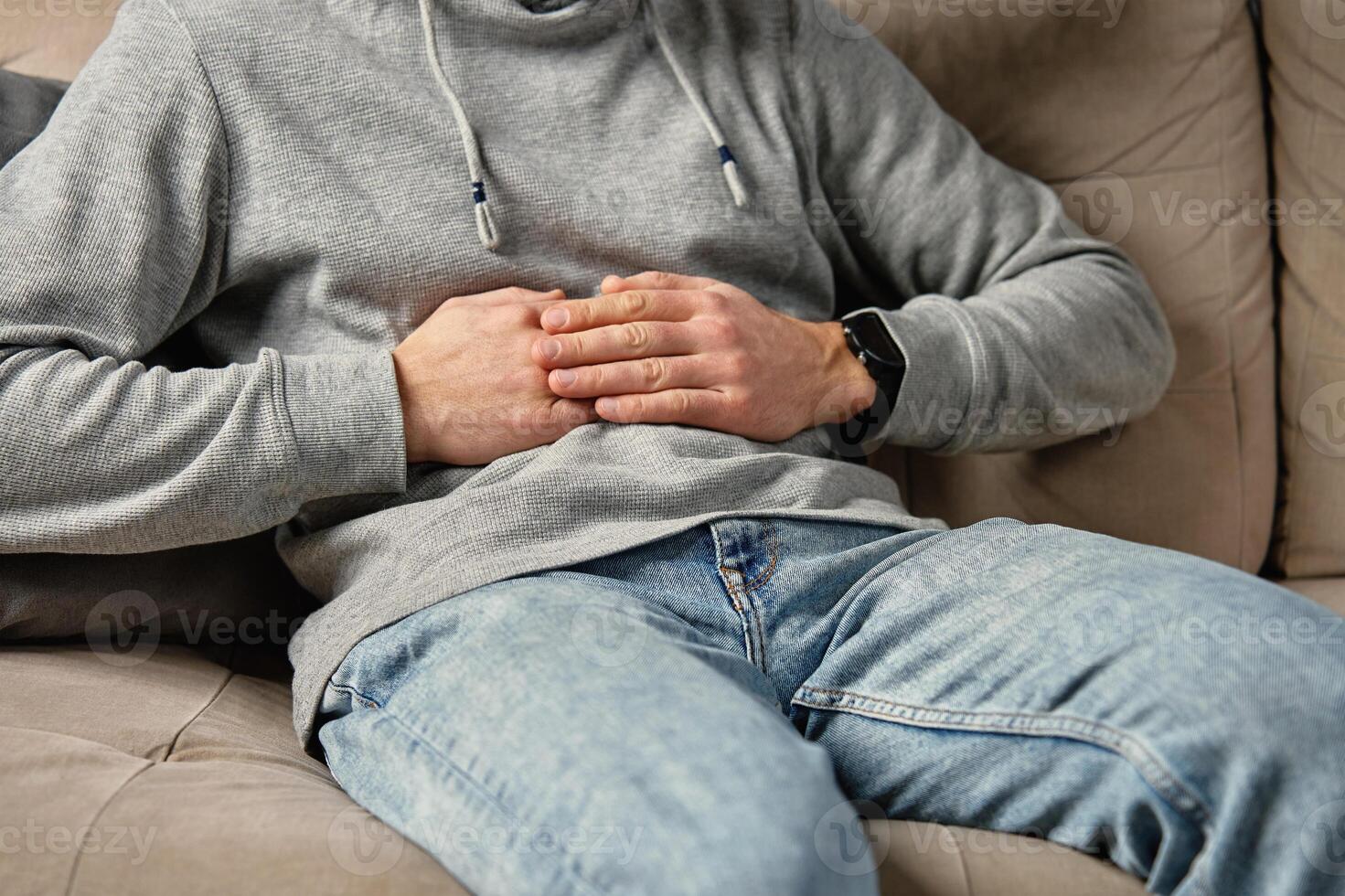 Close-up of man sitting on a couch at home, clutching his stomach, suffering from abdominal pain or cramps. Person suffering from health issues, digestion problems or food poisoning photo