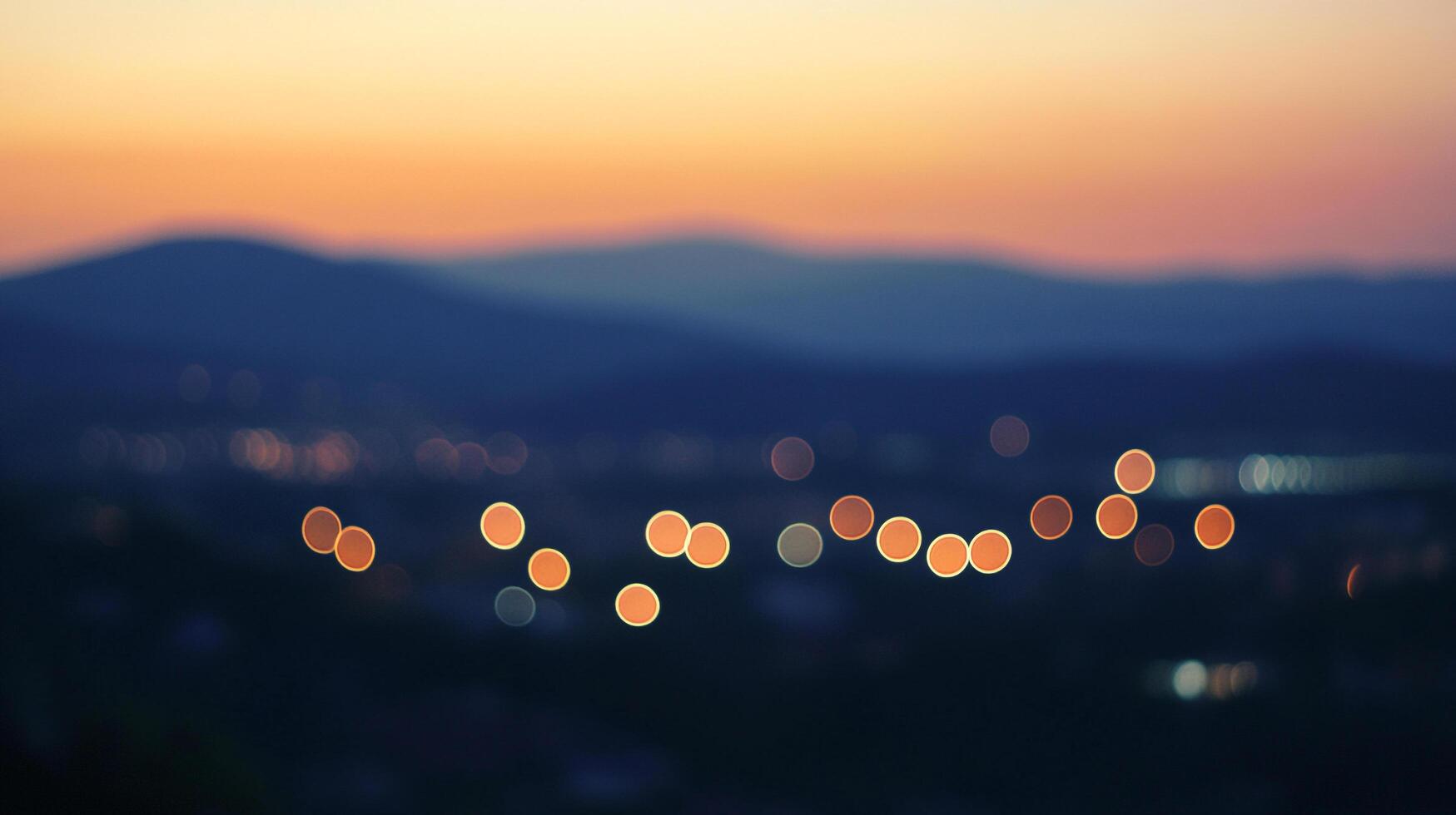 Distant city lights glow against a backdrop of mountains silhouetted by a colorful sunset, creating a dreamy and atmospheric scene. photo