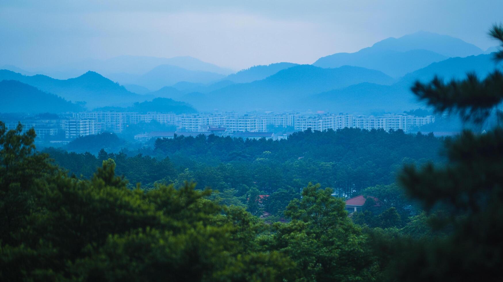 Landscape featuring lush greenery and a skyline of buildings set against a backdrop of hazy, blue-toned mountains on a serene, overcast day. photo